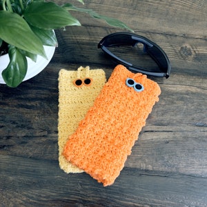 Op de afbeelding: Twee gebreide brillenkokers, een gele en een oranje, met blauwe en oranje knoopjesogen. De kokers liggen op een houten oppervlak.
