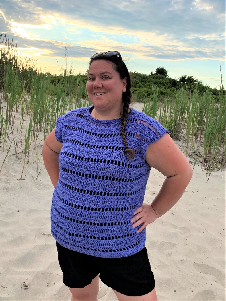 Puede incluir: Una mujer lleva una blusa de crochet morada de corte holgado y ligeramente recortada. La blusa tiene un patr&oacute;n de rayas horizontales con detalles calados. La mujer est&aacute; de pie en una playa de arena.