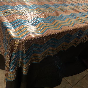 Könnte beinhalten: Eine Pailletten-Tischdecke mit Zickzackmuster in Gold, Türkis und Roségold. Die Tischdecke ist über einen Tisch drapiert, wobei am unteren Rand ein schwarzer Stoffrock zu sehen ist. Die Pailletten erzeugen einen schimmernden Effekt.