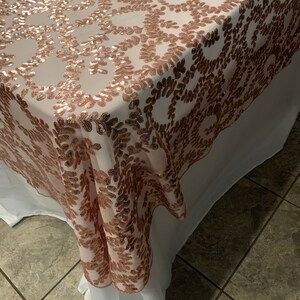 May include: A rose gold sequined tablecloth with a delicate floral pattern. The tablecloth is draped over a white table and has a subtle sheen.