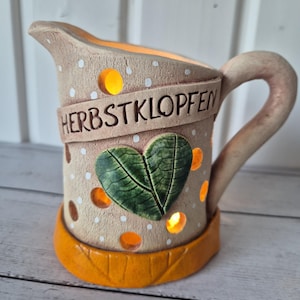 Könnte beinhalten: Eine beige, krugförmige Keramik-Laterne mit Henkel, weißen Punkten, einem grünen herzförmigen Blatt-Applikat und einem orangefarbenen Boden. Der Schriftzug HERBSTKLOPFEN ist auf einem Band eingeprägt, mit kreisförmigen Aussparungen für den Lichtschein.