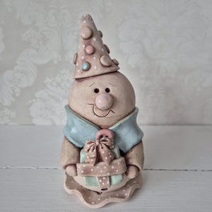 Könnte beinhalten: Eine skurrile Keramikfigur eines lächelnden Charakters mit einem gepunkteten Partyhut und einem hellblauen Kragen. Die Figur hält eine Geschenkbox mit einer Schleife. Die Farbpalette umfasst Beige, Rosa und Hellblau.
