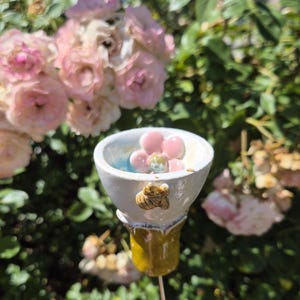 Könnte beinhalten: Handgefertigter Keramik-Gartenstecker mit einer weißen Schale, einem rosa Blumendesign und einem kleinen Bienenakzent. Der Stecker hat einen gelb-braunen Stiel, ideal, um jedem Garten oder Blumenbeet einen Hauch von Laune zu verleihen.