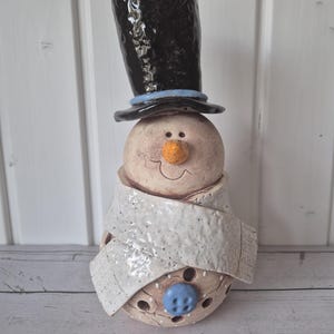 Könnte beinhalten: Eine Keramik-Schneemannfigur mit einem glänzenden schwarzen Zylinder mit hellblauem Band, einem strukturierten weißen Schal und einem einzelnen blauen Knopf am runden Körper. Der Schneemann hat eine kleine orangefarbene Nase.