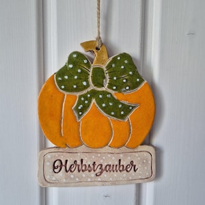 Könnte beinhalten: Ein hängendes Keramik-Kürbis-Ornament in leuchtendem Orange mit einer grünen Schleife mit weißen Punkten. Unter dem Kürbis befindet sich ein cremefarbenes, rechteckiges Schild mit der braunen Aufschrift "Herbstzauber", umrandet von weißen Punkten.