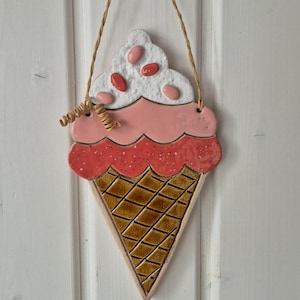 Könnte beinhalten: Ein keramischer Wandschmuck in Form einer Eiswaffel mit braunem Waffelmuster, einer gesprenkelten roten Kugel, einer glatten rosa Kugel und einer weißen, texturierten Spitze mit ovalen rosa Streuseln, aufgehängt an einer dünnen, braunen Kordel.