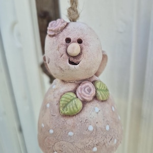 Könnte beinhalten: Handgefertigte Keramikglocke in hellem Rosa, verziert mit einem lächelnden Gesicht, einer Rose auf dem Kopf und floralen Akzenten. Die Glocke ist an einer natürlichen Schnur aufgehängt, ideal für die Heimdekoration.