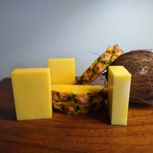 Peut inclure: Quatre barres de savon jaunes avec un ananas et une noix de coco au centre. L'ananas est tranché et la noix de coco est brune.