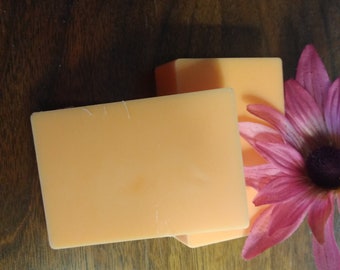 May include: Two rectangular bars of orange soap on a wooden surface. A pink flower is in the background.