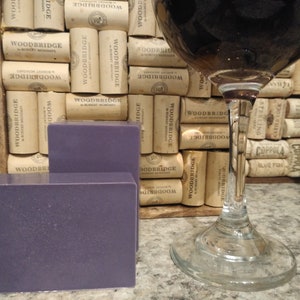 May include: Two bars of purple soap sit on a counter next to a glass of red wine. The wine glass is sitting on a background of cork wine bottle stoppers.