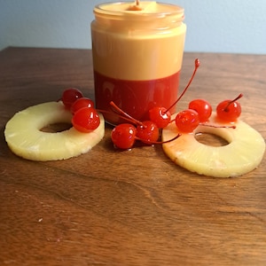 Può includere: Un barattolo di vetro con una candela a due strati, gialla e rossa. La candela è circondata da fette di ananas e ciliegie rosse.