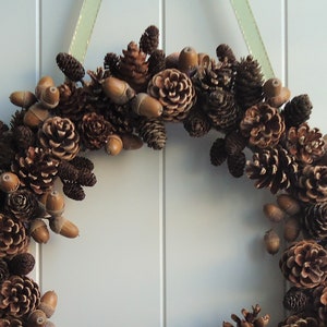 Puede incluir: Una corona de pi&ntilde;as marrones y beige con bellotas colgando de ella. La corona est&aacute; colgada en una pared blanca.