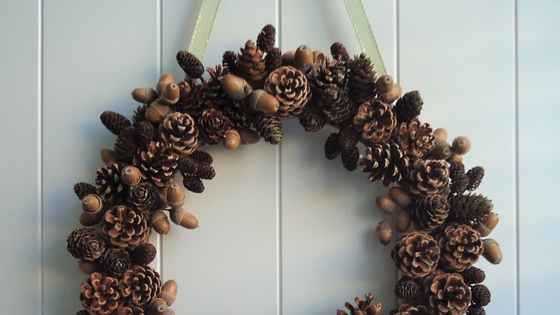 Puede incluir: Una corona de pi&ntilde;as marrones y beige con bellotas colgando de ella. La corona est&aacute; colgada en una pared blanca.
