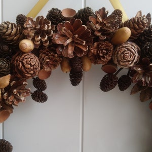 Puede incluir: Una corona de pi&ntilde;as marrones y beige con bellotas y otros elementos naturales.