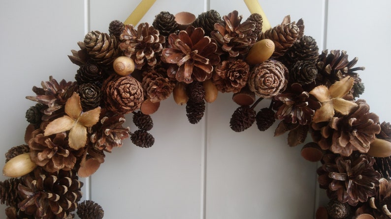 Puede incluir: Una corona de pi&ntilde;as marrones y beige con bellotas y otros elementos naturales.