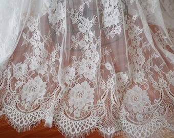 May include: Close-up of white lace fabric with a floral design. The lace features detailed patterns of flowers and leaves, with a scalloped edge. The delicate fabric is draped, showcasing its texture and design.