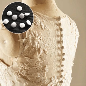 Puede incluir: Botones de plástico blanco para coser o manualidades. Los botones son pequeños y redondos, con la parte posterior plana y un centro elevado. Se muestran sobre un fondo negro.