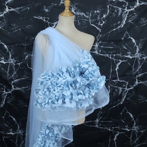 Puede incluir: Una tela de tul azul claro drapeada sobre un maniquí, adornada con racimos de flores de tela azul a juego. Las flores se concentran en el hombro y el borde inferior de la tela, creando un efecto decorativo. El fondo es un patrón de mármol blanco y negro.