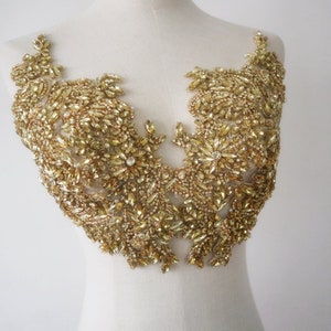 Könnte beinhalten: Goldgelbe, mit Perlen besetzte Applikation mit floralem Design. Die Applikation ist dicht mit kleinen, funkelnden Kristallen und größeren, facettierten Steinen bedeckt. Das Design ist symmetrisch, mit einem zentralen Blumenmotiv und zarten, blattartigen Mustern.