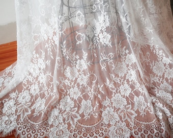 May include: White lace fabric with a floral pattern. The lace features a scalloped edge and is draped over a wooden surface. The intricate design includes various flower and leaf motifs, creating a delicate and elegant texture.