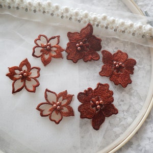 Può includere: Sei applicazioni di fiori in pizzo marrone con decorazioni perlate. Le applicazioni sono disposte in cerchio su uno sfondo bianco.