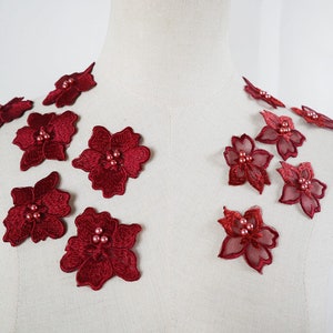 May include: Burgundy floral appliques with embroidered details and pearl-like beads. The appliques are displayed on a mannequin, suggesting their use in fashion or craft projects. The flowers are a deep red colour.