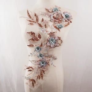 May include: A floral applique with a delicate design. The applique features 3D flowers in shades of dusty rose, light blue, and copper, embellished with sequins and beads. The design includes leaves and stems, all on a sheer, off-white mesh backing.