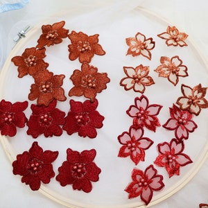 Pode incluir: Conjunto de aplicações de flores bordadas em vermelho e laranja com detalhes em pérolas. As aplicações estão dispostas sobre um fundo branco.
