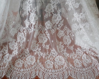 May include: Ivory lace fabric with a floral pattern. The lace features detailed flower and leaf designs, with a scalloped edge. The fabric is draped, showing the intricate details and texture of the lace. Ideal for bridal wear or other delicate projects.