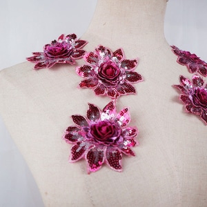 May include: A selection of pink and burgundy sequined floral appliques. The appliques are displayed on a mannequin, highlighting their design. Each flower has a central rose and petal-shaped sequins.