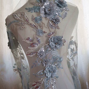 Könnte beinhalten: Eine zarte silberne und graue Blumenapplikation mit Pailletten und Perlen. Die Applikation befindet sich auf einem durchsichtigen Netzstoff und wäre perfekt, um einem Brautkleid oder einem anderen Kleidungsstück für besondere Anlässe einen Hauch von Eleganz zu verleihen.