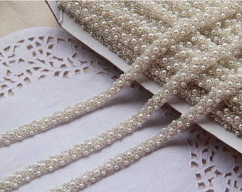 May include: White pearl trim with a beaded design. The trim is made of small, white pearls that are sewn onto a clear, thin ribbon. The trim is perfect for adding a touch of elegance to any project.