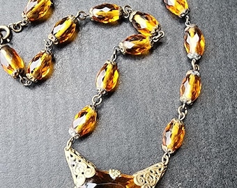 May include: Vintage amber-colored necklace featuring faceted oval beads and a large, geometric pendant. The necklace has a delicate chain and ornate metalwork, creating an elegant and classic look. The pendant is a rich, warm amber color.