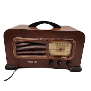 May include: Vintage Philco radio in a dark brown wooden cabinet. The radio features a speaker grill, a dial with numbers, and three control knobs. The word "PHILCO" is printed on the front. A black handle is on top.