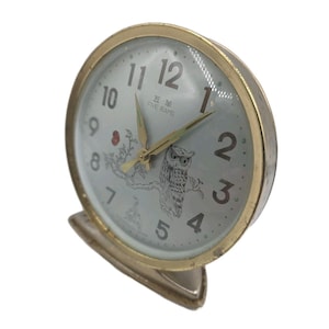 May include: Vintage gold-tone alarm clock with a light blue face. The clock features black numerals, gold hands, and an owl illustration. The text "FIVE RAMS" is printed on the clock face. The clock has a stand and a gold-tone bezel.