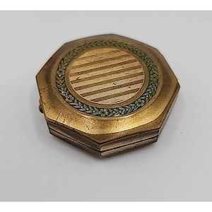 May include: Vintage octagonal compact with a gold-tone finish. The center features a circular design with horizontal lines, surrounded by a green floral border. The compact is closed and shows signs of age.