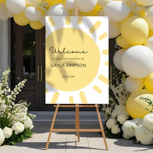 May include: A baby shower welcome sign with a yellow sun design, displaying the text "Welcome to the baby shower of Layla Simpson" and the date 01.01.2026. The sign is on a wooden easel, with white and yellow balloons and floral decorations.