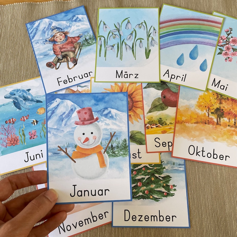 May include: A set of 12 colorful cards featuring illustrations of different months of the year. Each card has a month name written in German and a corresponding image. For example, the January card shows a snowman, the February card shows a person sledding, and the March card shows snowdrops.