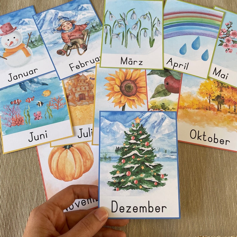 May include: A set of 12 colorful cards featuring illustrations of different months of the year. Each card has a month name written in German and a corresponding image. For example, the January card shows a snowman, the March card shows snowdrops, and the December card shows a Christmas tree.