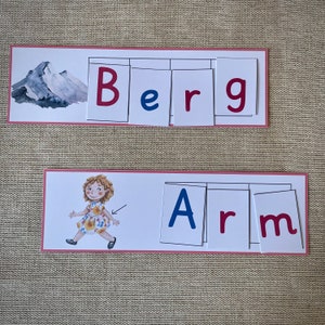 May include: Two sets of flashcards with pink borders, each set spelling out a word. The first set spells out "Berg" with blue and red letters. The second set spells out "Arm" with blue and red letters. A cartoon illustration of a girl wearing a floral dress is on the second set of flashcards.