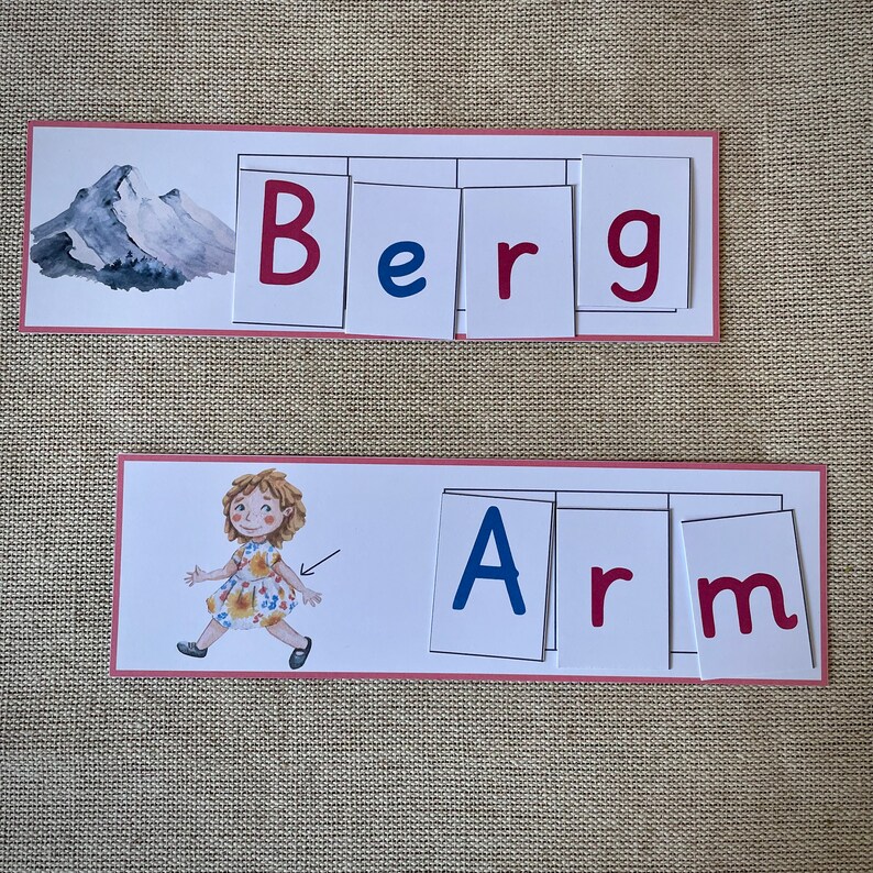 May include: Two sets of flashcards with pink borders, each set spelling out a word. The first set spells out "Berg" with blue and red letters. The second set spells out "Arm" with blue and red letters. A cartoon illustration of a girl wearing a floral dress is on the second set of flashcards.