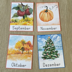 May include: Four watercolor illustrations on cards depicting the months of September, October, November, and December. September shows red apples on a branch, October shows a group of trees with fall foliage, November shows a pumpkin, and December shows a Christmas tree with snow.