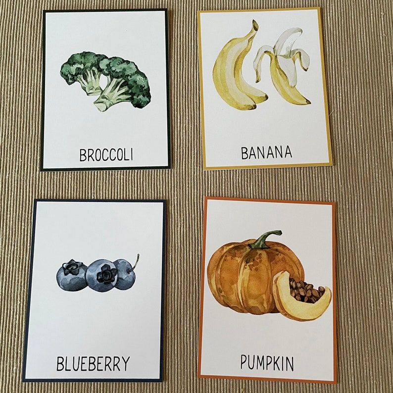 May include: Four illustrated cards with different fruits and vegetables. The cards are white with black borders. The first card shows a green broccoli with the word "Broccoli" printed below. The second card shows a yellow banana with the word "Banana" printed below. The third card shows three blue blueberries with the word "Blueberry" printed below. The fourth card shows an orange pumpkin with the word "Pumpkin" printed below.