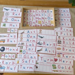 May include: A set of colourful flashcards with German words and pictures. The words include "Wal", "Rad", "Oma", "Hut", "Zug", "Bus", "Mond", "Korb", "Huf", "Ast", "Opa", "Arm", "Lok", "Tor", "Glas", "Saft", "Turm", "Gras", "Holz", "Gans", "Salz", "Ring", "Pilz", "Kind", "Bild", "Brot", "Kern", "Herz", "Gold", and "Berg".