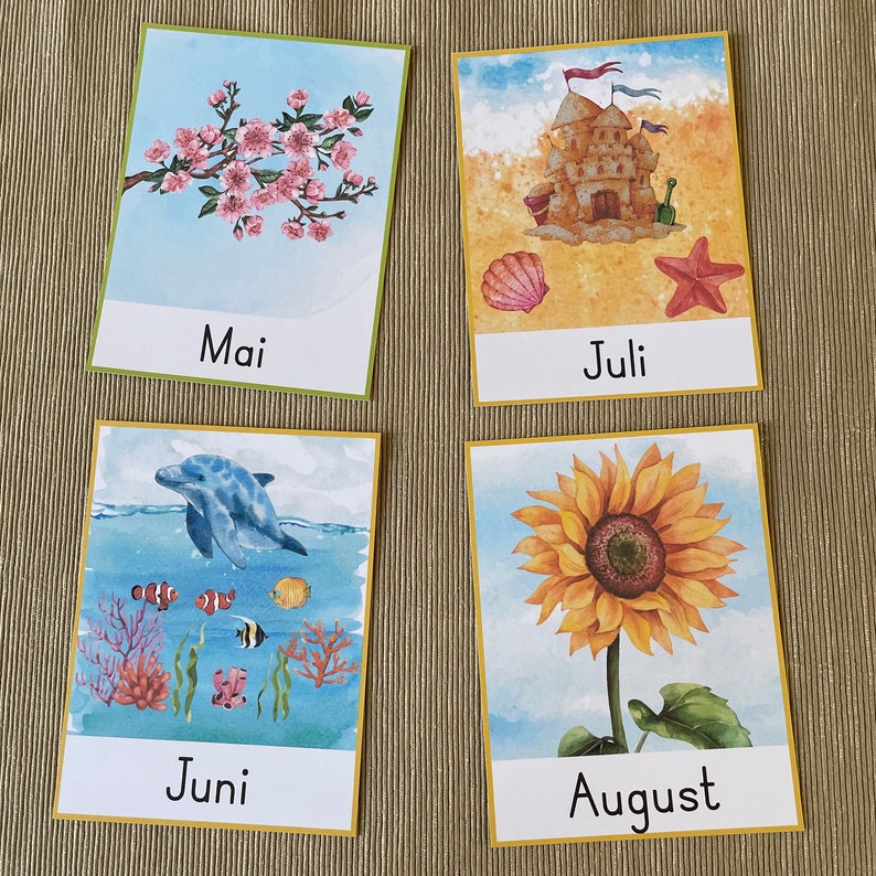 May include: Four illustrated cards with the months of May, June, July, and August. The May card features a pink cherry blossom tree. The June card features a blue dolphin swimming in the ocean with colorful fish. The July card features a sandcastle with a red starfish and a seashell. The August card features a yellow sunflower.