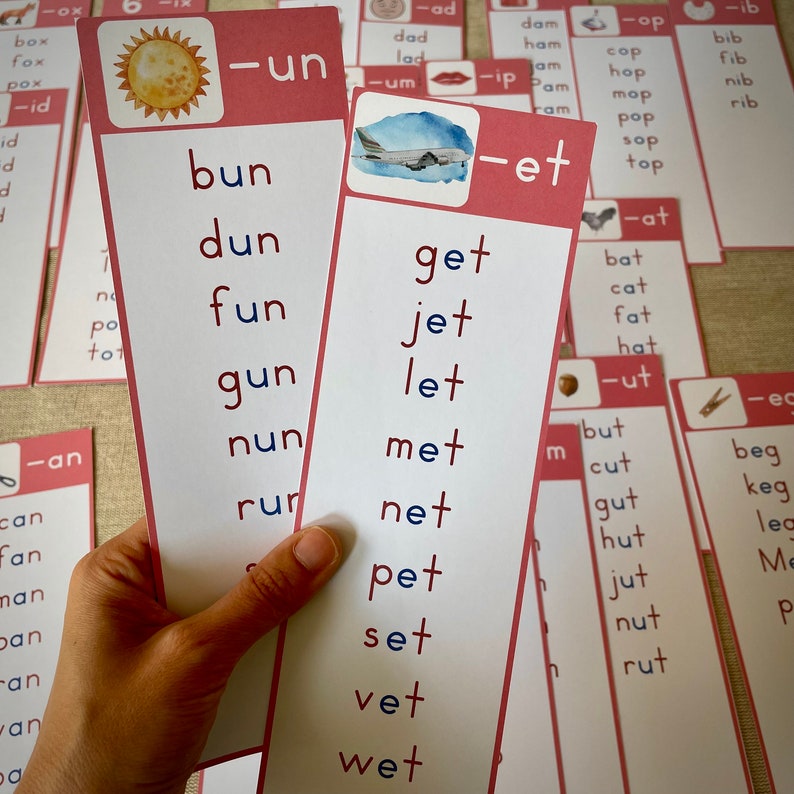 May include: Two pink and white flashcards with the words "bun, dun, fun, gun, nun, run, sun" and "get, jet, let, met, net, pet, set, vet, wet" written in blue and red. The flashcards are held in a hand.