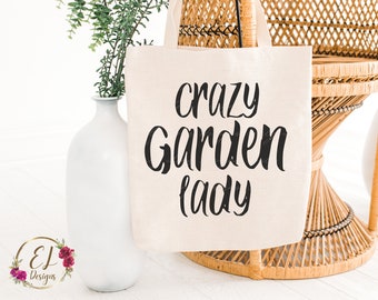 Op de afbeelding: Een witte canvas draagtas met zwarte tekst die "crazy garden lady" zegt. De tas ligt op een rieten stoel.