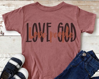 Op de afbeelding: Een maronkleurig T-shirt met korte mouwen met de tekst "LOVE GOD love people" in zwarte en oranje letters. Het shirt ligt op een houten oppervlak met een paar blauw-witte sneakers en een blauwe jeans.