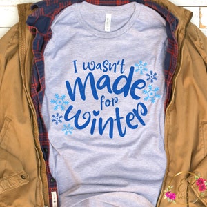 May include: A light gray t-shirt with the text "I wasn't Made for Winter" in blue script, surrounded by blue snowflakes. The shirt is displayed on a wooden surface, layered with a plaid shirt and a brown jacket.