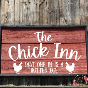 以下が含まれることがあります： 黒い縁取りのある赤い木製の看板に、白い筆記体で「The Chick Inn」と書かれています。タイトルの下には、白い文字で「Last one in is a rotten egg」と書かれています。テキストの両側に、白い鶏のシルエットが2つあります。